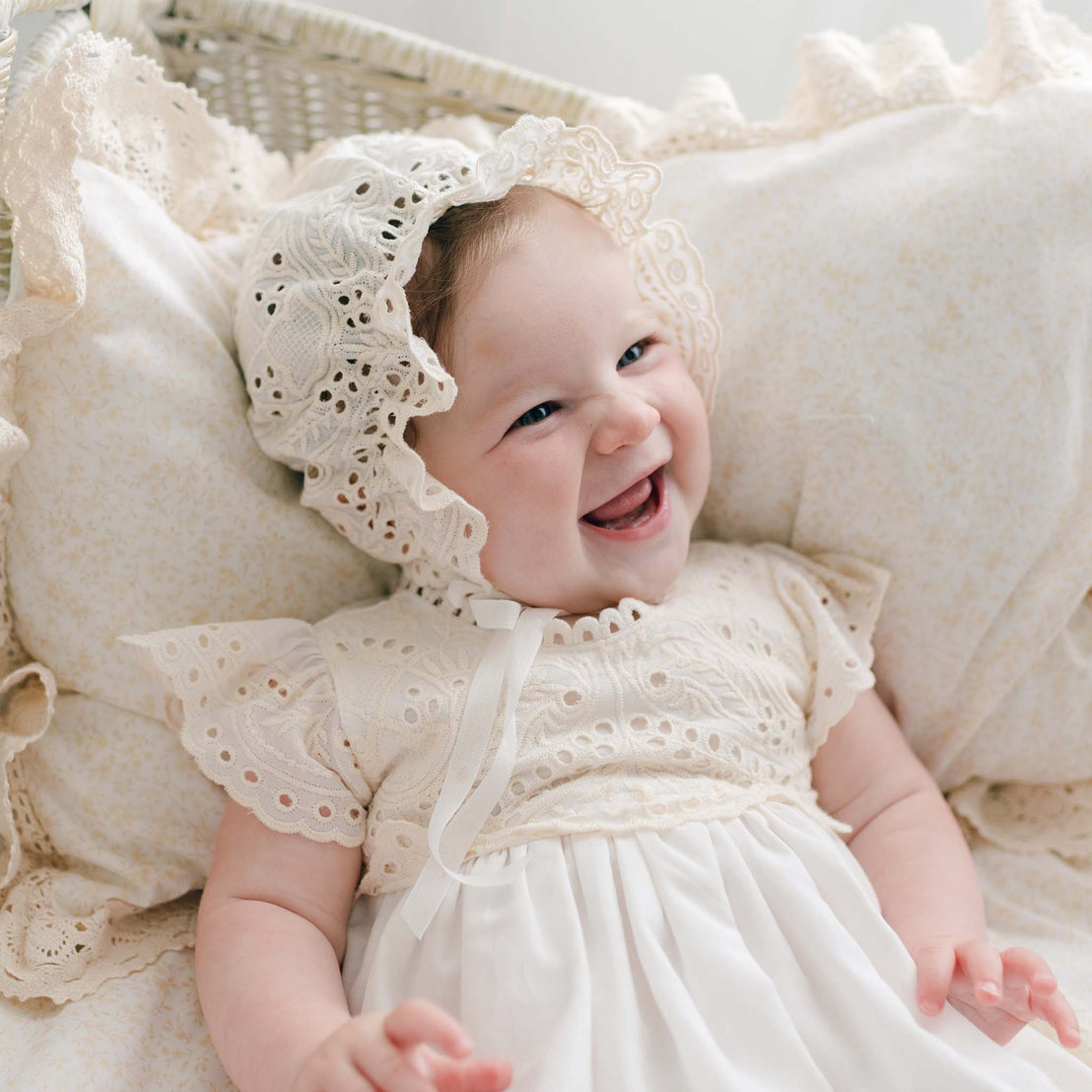 A baby is lying on a pillow, wearing the Ingrid Tiered Gown & Bonnet with beautiful lace details. The baby is smiling brightly with visible dimples, creating a joyful expression. The background includes a lace-trimmed pillow and a woven basket.