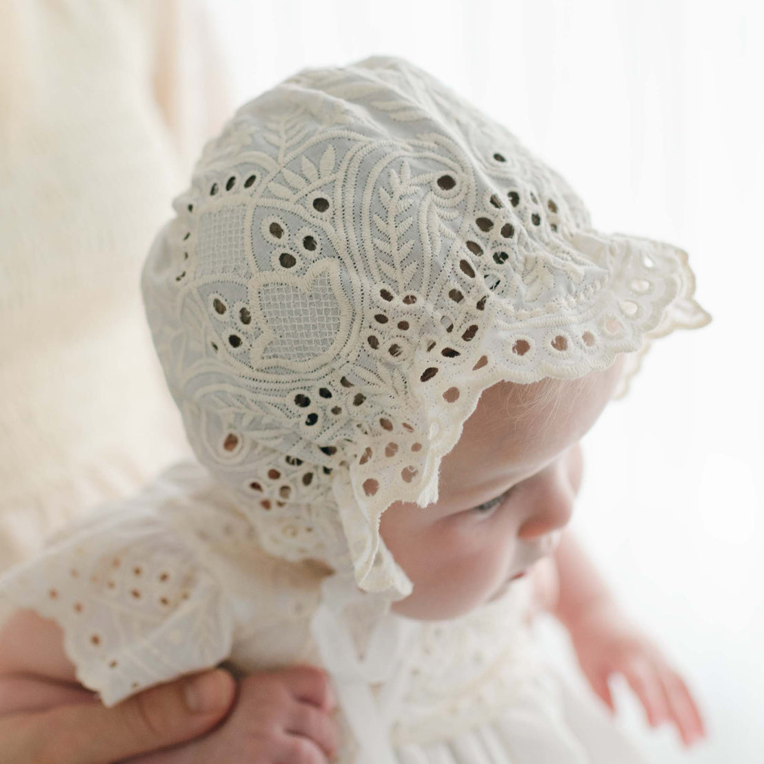 An adult holds a baby wearing an intricately patterned bonnet and a matching outfit. The white bonnet, part of the lovely Ingrid Tiered Gown & Bonnet handmade in the USA, features lace and embroidery details. The baby is looking slightly downward and to the side. The background is softly lit, emphasizing the delicate attire.