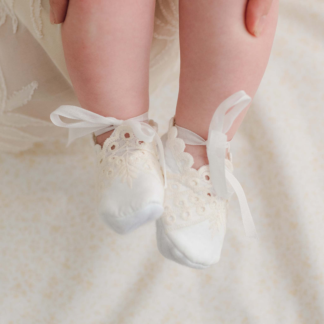 A close-up of a baby's feet wearing Ingrid Booties, adorned with vintage lace trim and ribbon ties. The baby's legs are being gently held by an adult against a backdrop of cream-colored fabric with a subtle pattern.