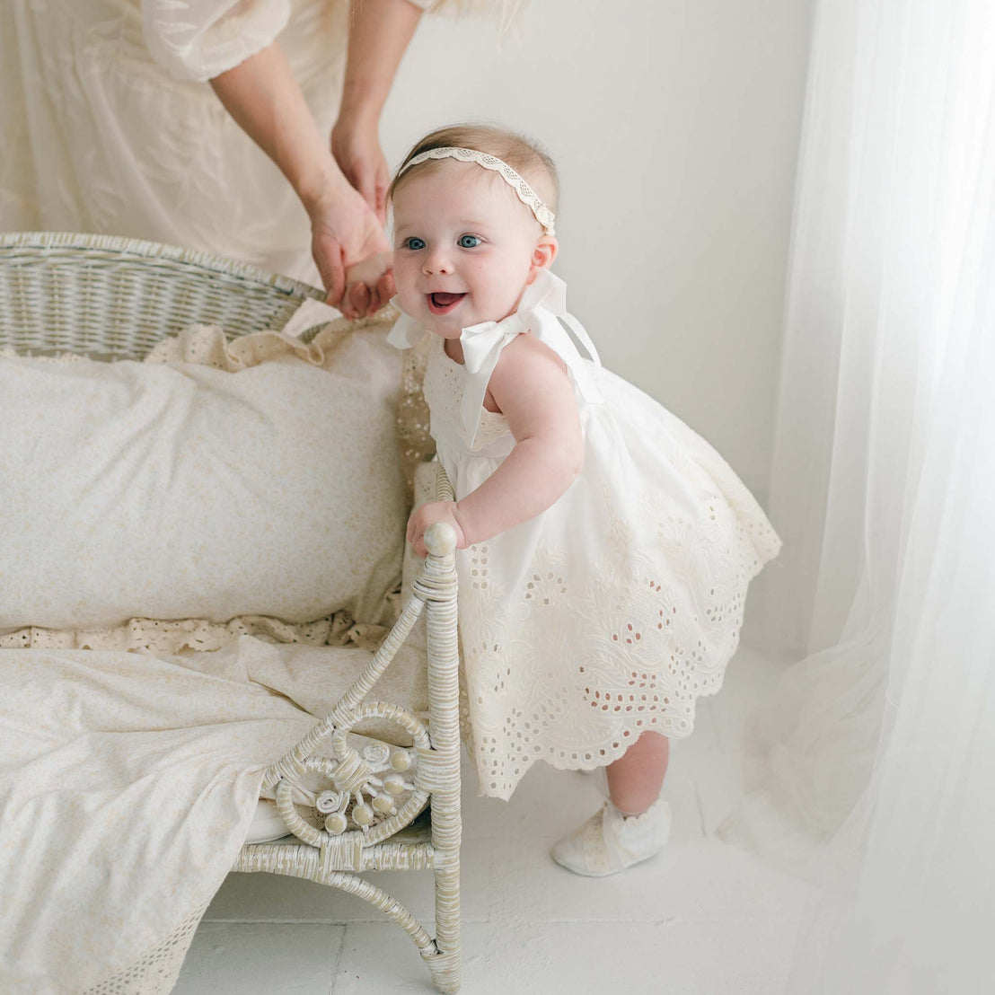 A baby dressed in an Ingrid Romper Dress, paired with a headband, stands holding onto a wicker crib while an adult's hands provide gentle support. The softly lit room features white decor.