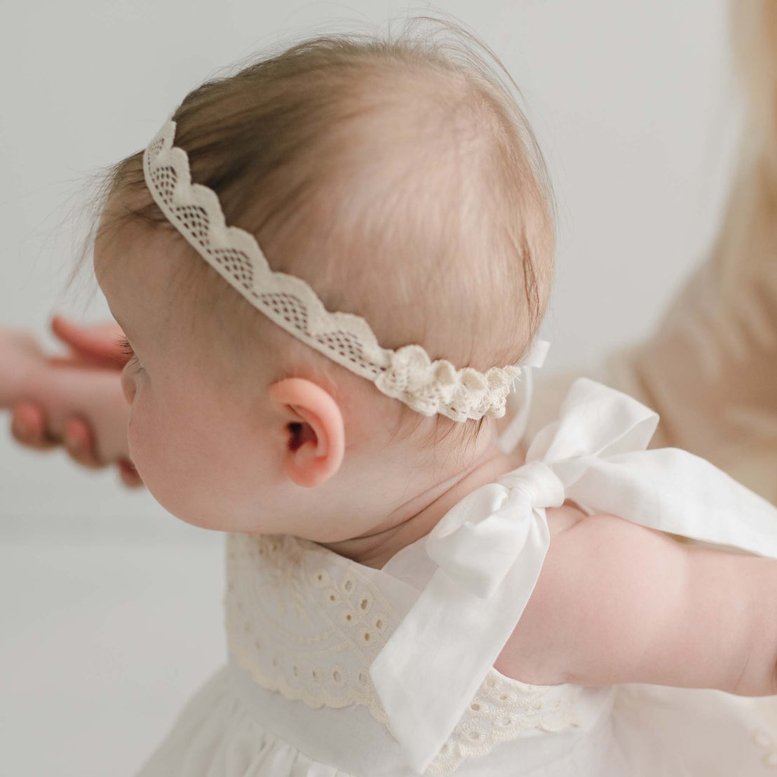 A baby in a delicate Christening Gown with an Ingrid Lace Headband and a large bow on the shoulder is held by an adult's hand. The baby, viewed in profile and facing left, stands out against the softly blurred background.