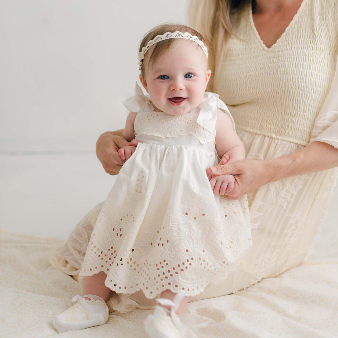 A baby in a white Ingrid Romper Dress with lace details, a headband, and white shoes sits on a woman's lap. The woman is wearing a light-colored dress, and both are seated on a light fabric surface. The baby smiles at the camera.