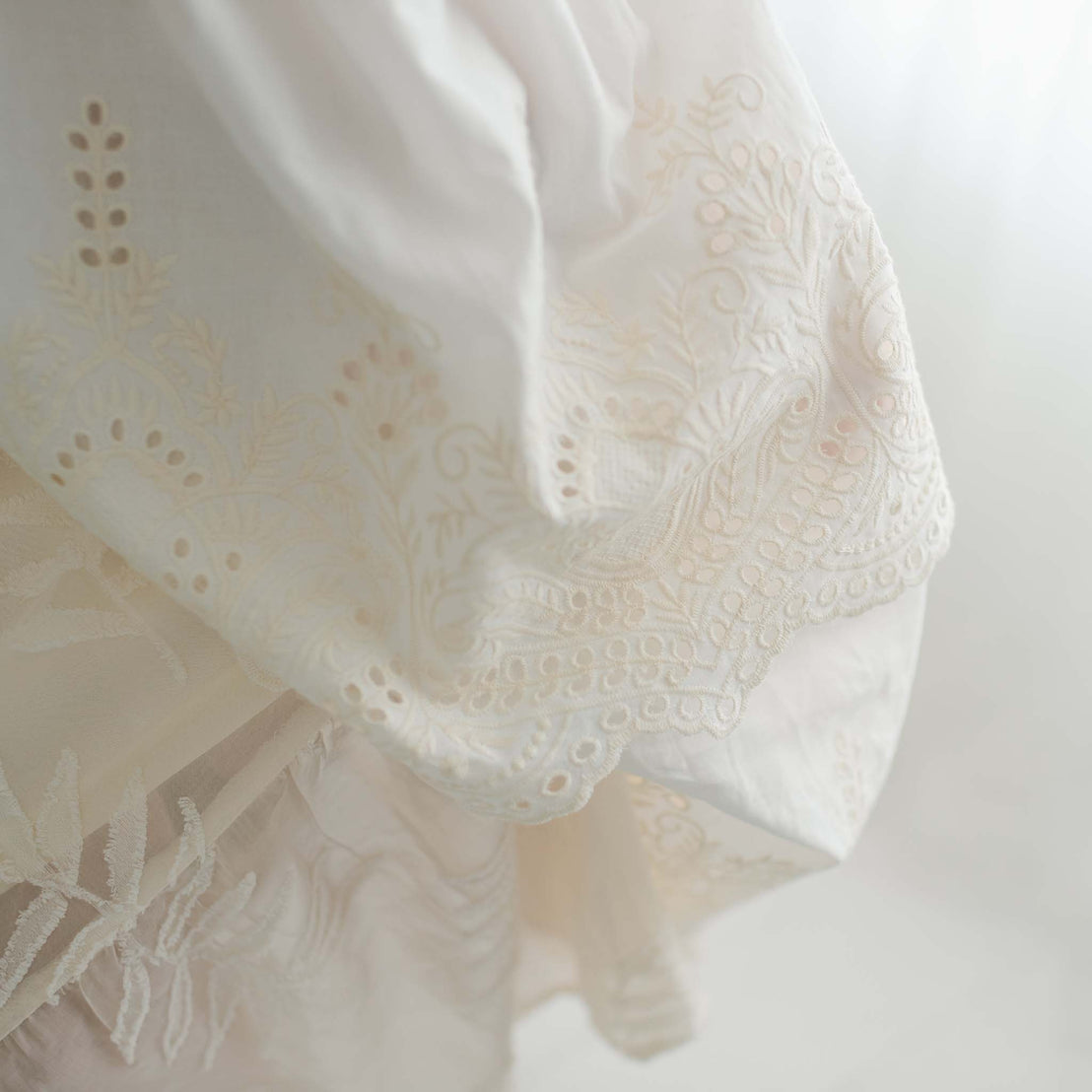 Close-up of the Ingrid Tiered Gown & Bonnet, an elegant piece featuring intricate embroidery and lace detailing, perfect for a handmade girls' gown. The fabric appears slightly layered, with soft light illuminating its delicate patterns and textures, making it ideal for a baby blessing gown or christening gown.