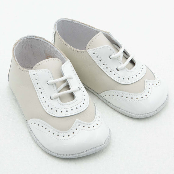 A pair of Boys Ivory Two Tone Wingtip Shoes with laces is displayed on a light background. These handmade baby shoes feature a brogue design with perforated detailing and white soles. They are beautifully crafted from leather.