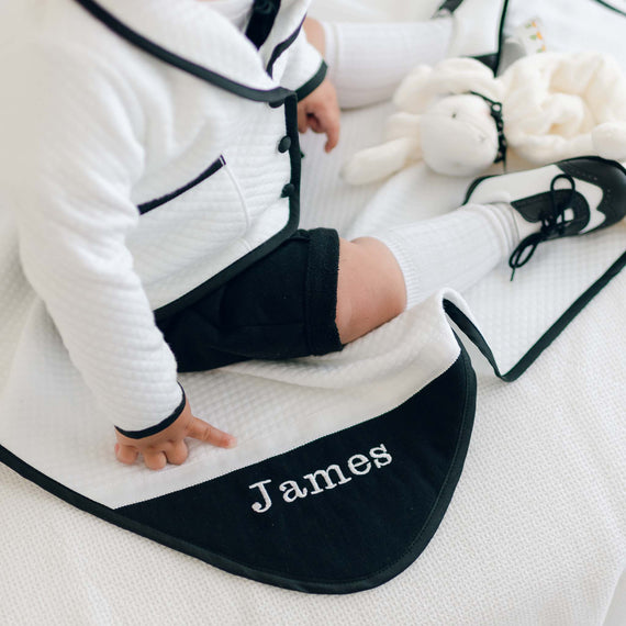 A baby dressed in a black and white outfit sits on the James Personalized Blanket, which is white and made of 100% cotton, with the name "James" embroidered on it. The personalized baby gift also includes matching black and white shoes and a plush toy placed beside them.