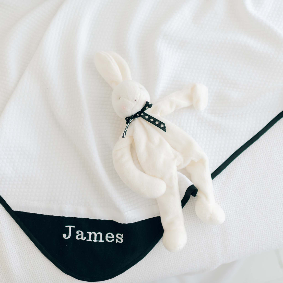 The James Personalized Blanket, crafted from 100% cotton and featuring "James" embroidered in blue on the corner, is beautifully spread out. Resting on the blanket is a plush white bunny adorned with a black ribbon around its neck, making it an ideal personalized gift for a baby.