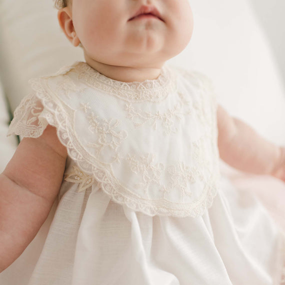 A close-up of a baby wearing a white, lace-trimmed dress and a Jessica Cotton Christening Bib. The baby’s face is partially visible, showing their chubby cheeks and closed lips. The dress features detailed floral embroidery and cap sleeves. The background is softly blurred, focusing on the baby.