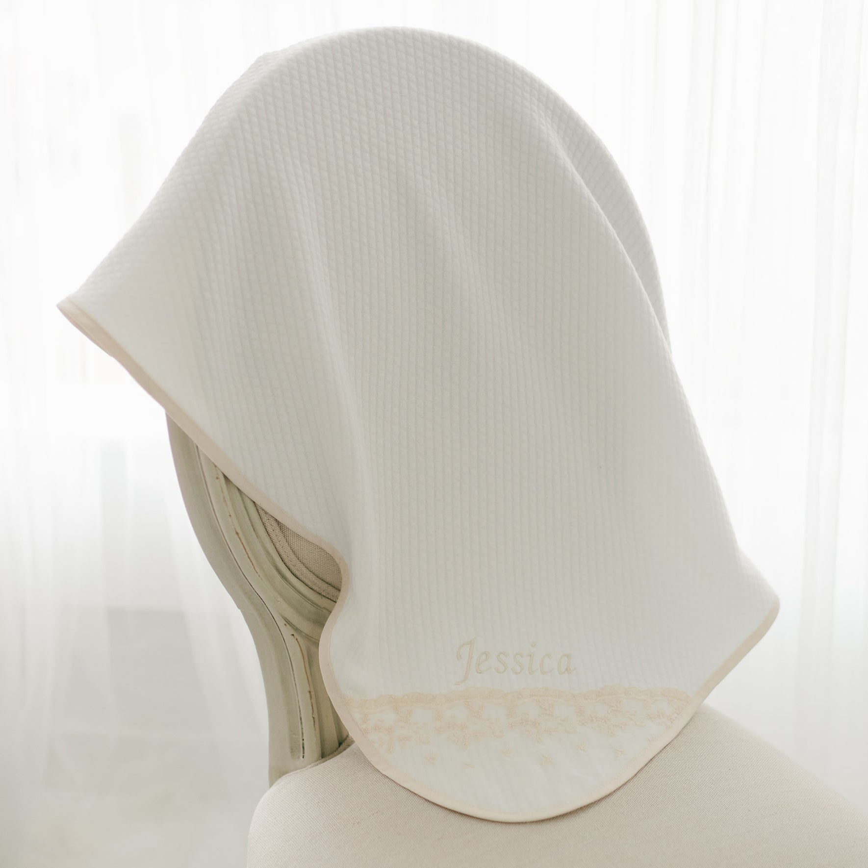 A white, handmade Jessica Personalized Blanket embroidered with the name "Jessica" is draped over the back of a chair. This Jessica Personalized Blanket has a textured surface and light brown trimming along the edge. The background is softly lit with white curtains, making it an elegant baby gift.