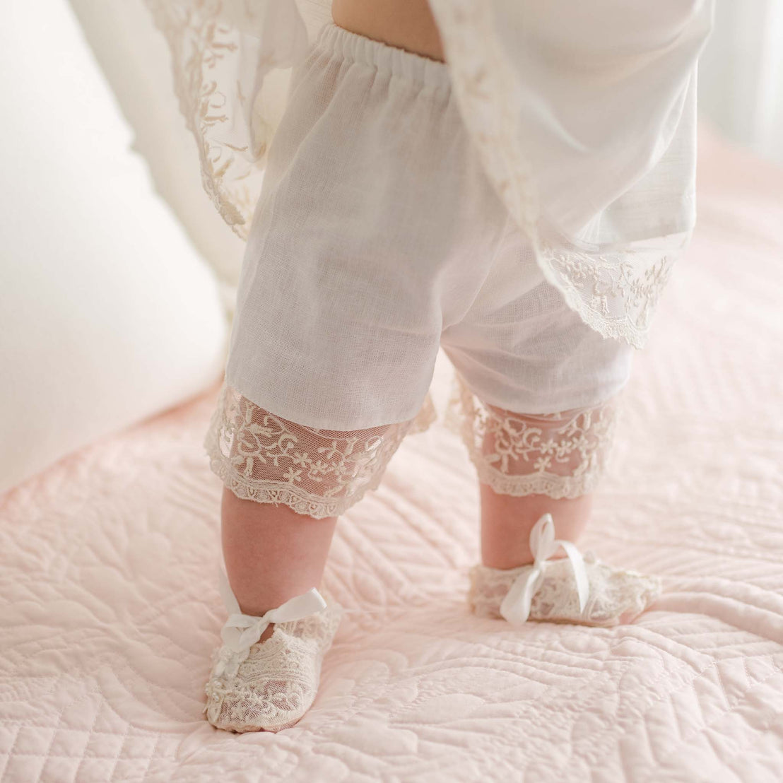 A baby stands on a pink quilted blanket, showcasing a vintage touch with white lace-trimmed bloomers and matching lace booties. The delicate details of the outfit perfectly complement the Jessica Linen Dress that gracefully drapes over part of the baby's upper body.