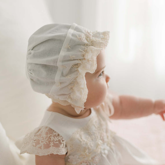 A baby wearing the Jessica Linen Bonnet looks to the side. The baby has a calm expression and is seated, with one hand slightly extended. The background is softly lit and white.