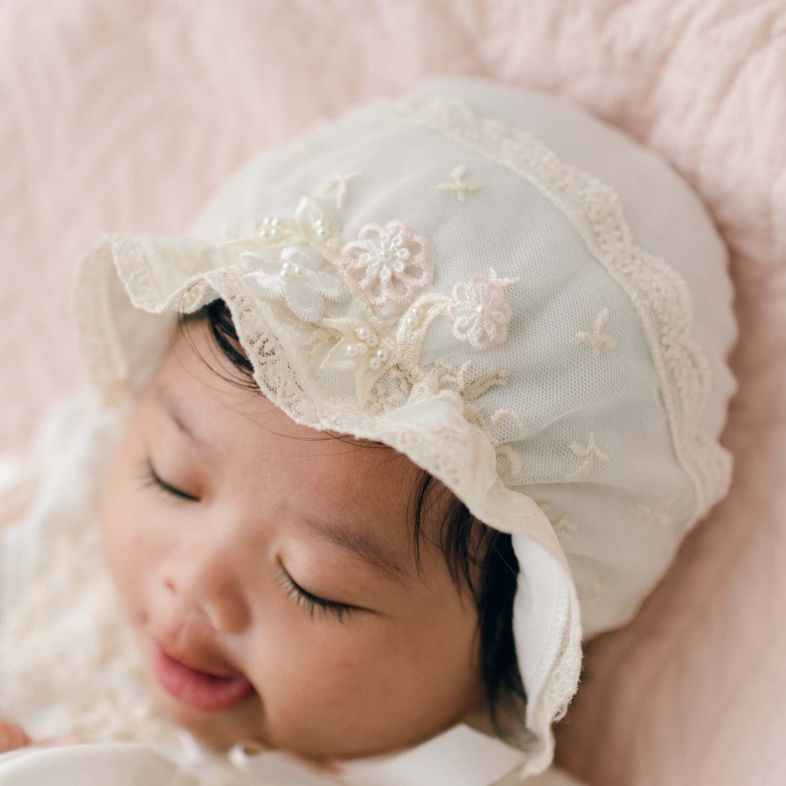 A close-up of a baby wearing the Jessica Linen Bonnet decorated with delicate floral appliqué. The baby has closed eyes and a slight smile. The background is soft and light-colored, complementing the baby's attire.