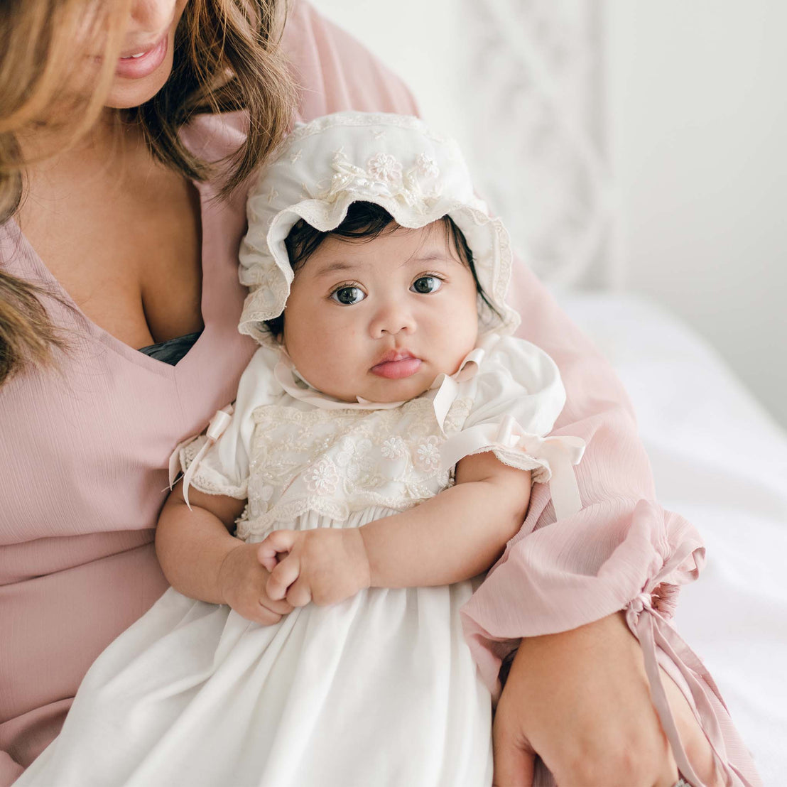 A baby dressed in a Jessica Newborn Gown & Bonnet sits on her mother's lap. The mother is wearing a pink blouse. The scene appears to be indoors, with a light and airy atmosphere. The baby looks directly at the camera with wide eyes.