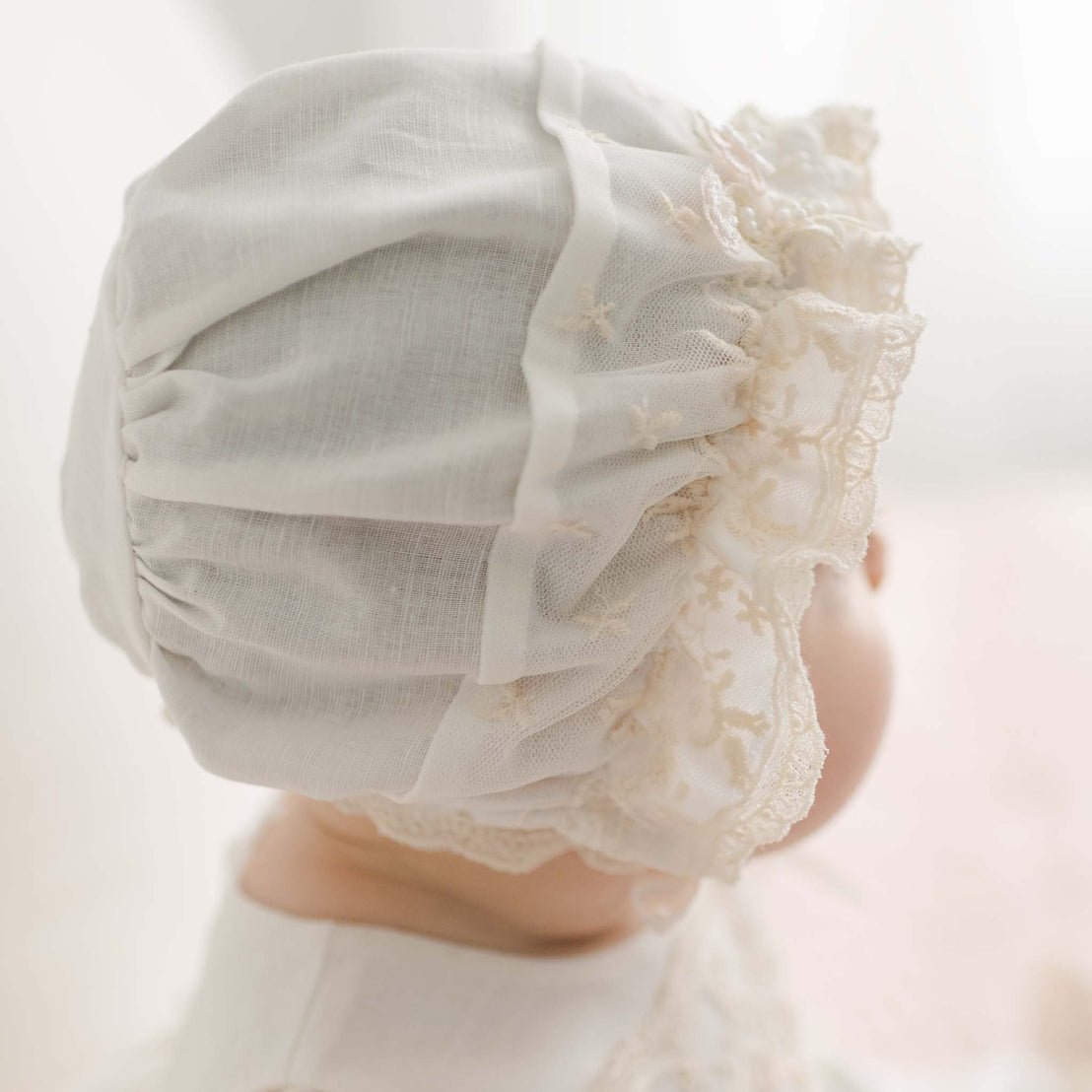 A close-up image of the back of a baby's head, wearing the Jessica Linen Bonnet. The bonnet is crafted with a linen/cotton blend in light ivory with a champagne lace detailing. The baby is facing away from the camera.