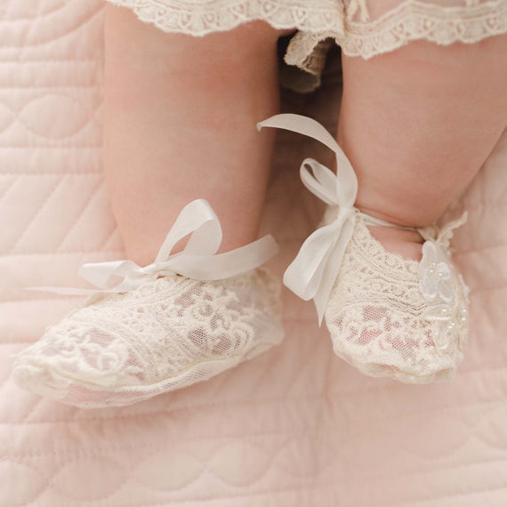Close-up image of a baby's feet wearing the Jessica Lace Booties with ribbon ties. The baby is lying on a quilted pink blanket.