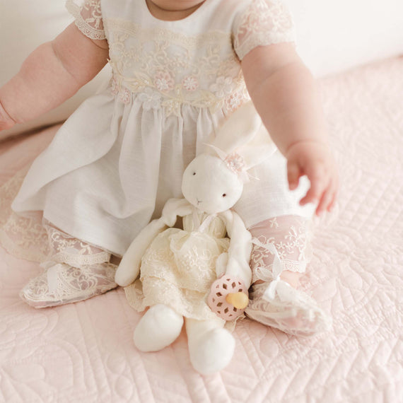 A baby wearing a white lace dress sits on a pink quilted blanket, holding a stuffed white bunny dressed in lace. The baby's arm is reaching outward, revealing detailed lace patterns on the dress and blanket, while a **Jessica Silly Bunny Buddy | Pacifier Holder** dangles nearby.