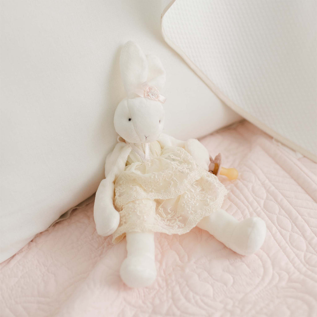 A Jessica Silly Bunny Buddy | Pacifier Holder, with a pink flower on its head and a cream lace dress, sits propped against white pillows on a pale pink quilted blanket.