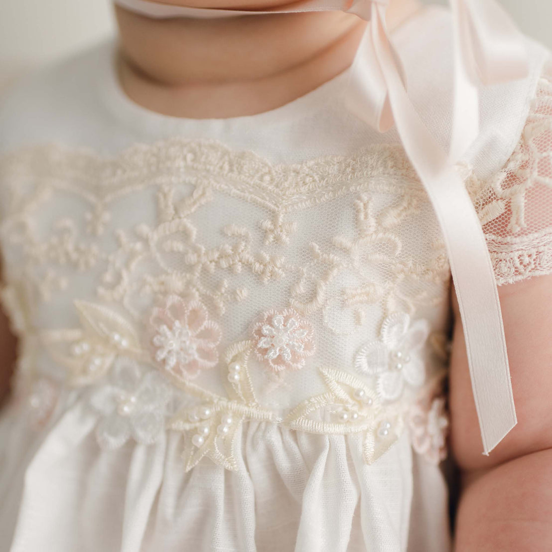 A close-up of a baby wearing the Jessica Linen Dress & Bloomers, showcasing intricate lace and pearl details. The fabric is adorned with floral embroidery that adds a vintage touch, complemented by a pink ribbon tied around the shoulder. The baby's face is not visible.