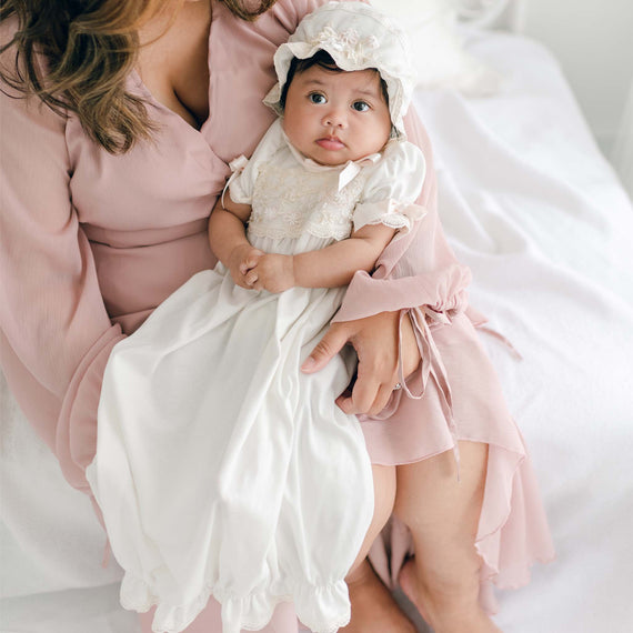 An infant wearing the Jessica Newborn Gown & Bonnet with delicate floral appliqués sits on the lap of an adult in a pink dress. The adult's arms are around the child, and they are seated on a light-colored surface. The setting appears soft and serene.