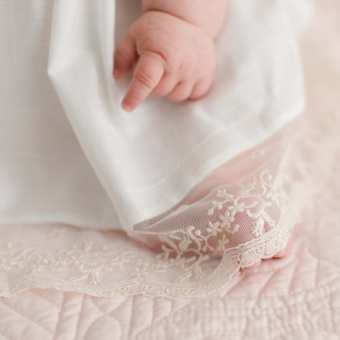 A baby's hand and foot are partially visible, adorned in the delicate Jessica Linen Dress & Bloomers with lace trim. The fabric gently drapes over a pink quilted surface, imbuing the scene with a vintage touch of softness and delicacy.