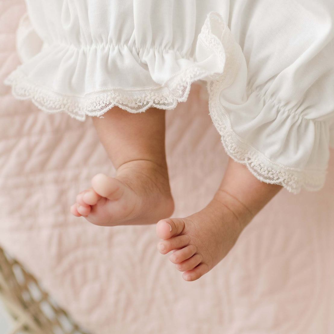 A close-up of a baby's legs and feet in the Jessica Newborn Gown. The baby is lying on a soft, quilted surface. The image focuses on the edges of the gown, highlighting the delicate lace trimming.