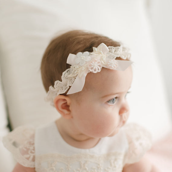 A baby wearing a Jessica Headband adorned with flowers and a bow, looking to the right. The baby is dressed in a white outfit with champagne lace detailing, resembling a christening gown. The background is soft-focused and light-colored.