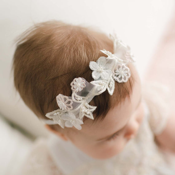 A close-up of a baby wearing a white lace headband adorned with embroidered flowers. The Jessica Beaded Flower Headband complements the baby's light brown hair and soft, light-colored outfit. The background is blurred, drawing attention to the baby and the Jessica Beaded Flower Headband.