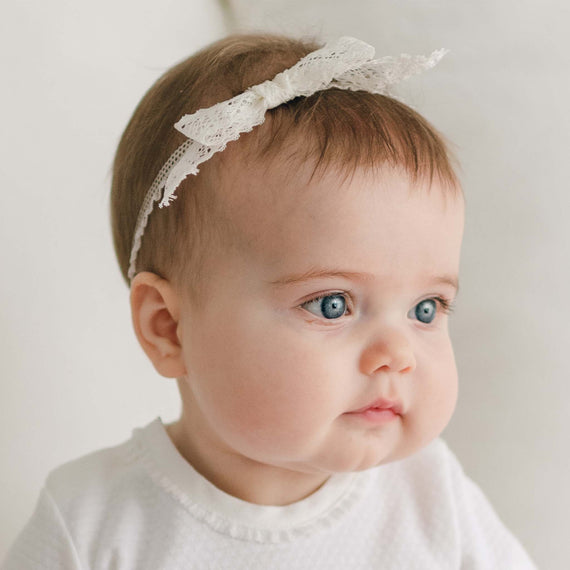 A baby with blue eyes and brown hair wearing a Hailey Lace Headband adorned with a delicate lace bow. The baby is dressed in a white outfit and looks to the side against a plain, light-colored background.