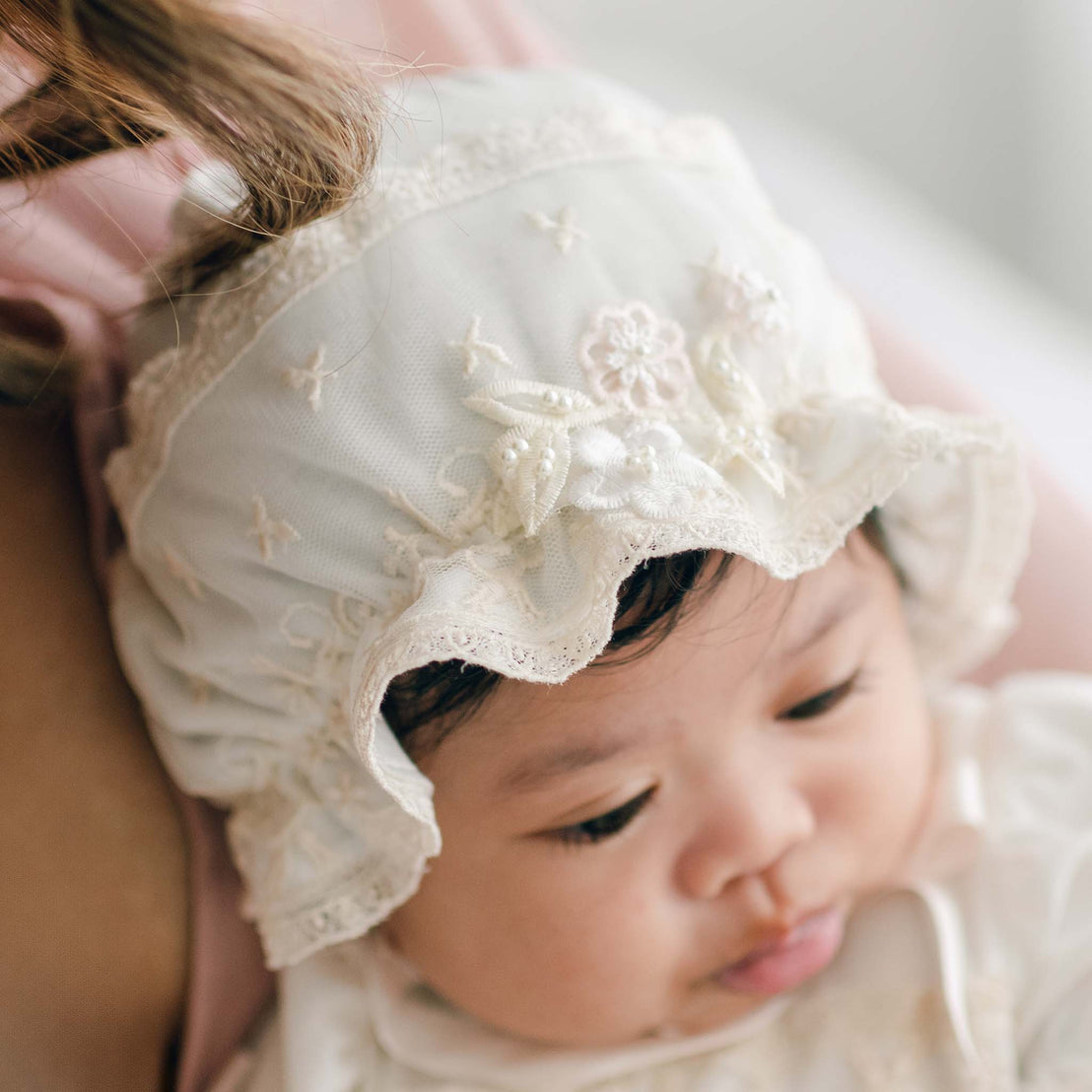 A close-up photograph of a baby wearing the Jessica Linen Bonnet. The baby has a neutral expression and is being cradled by an adult.