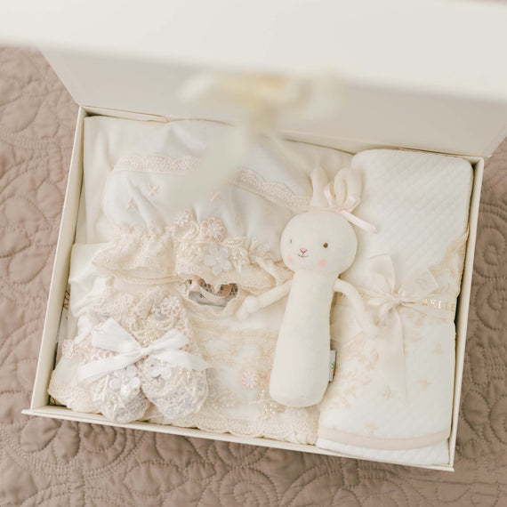 A white gift box filled with baby items, including crocheted booties with ribbon bows, a stuffed bunny toy, a handmade in the USA white blanket with a subtle pattern, and white baby clothing adorned with lace and floral details, all arranged neatly inside the Jessica Newborn Gift Set - Save 10%.