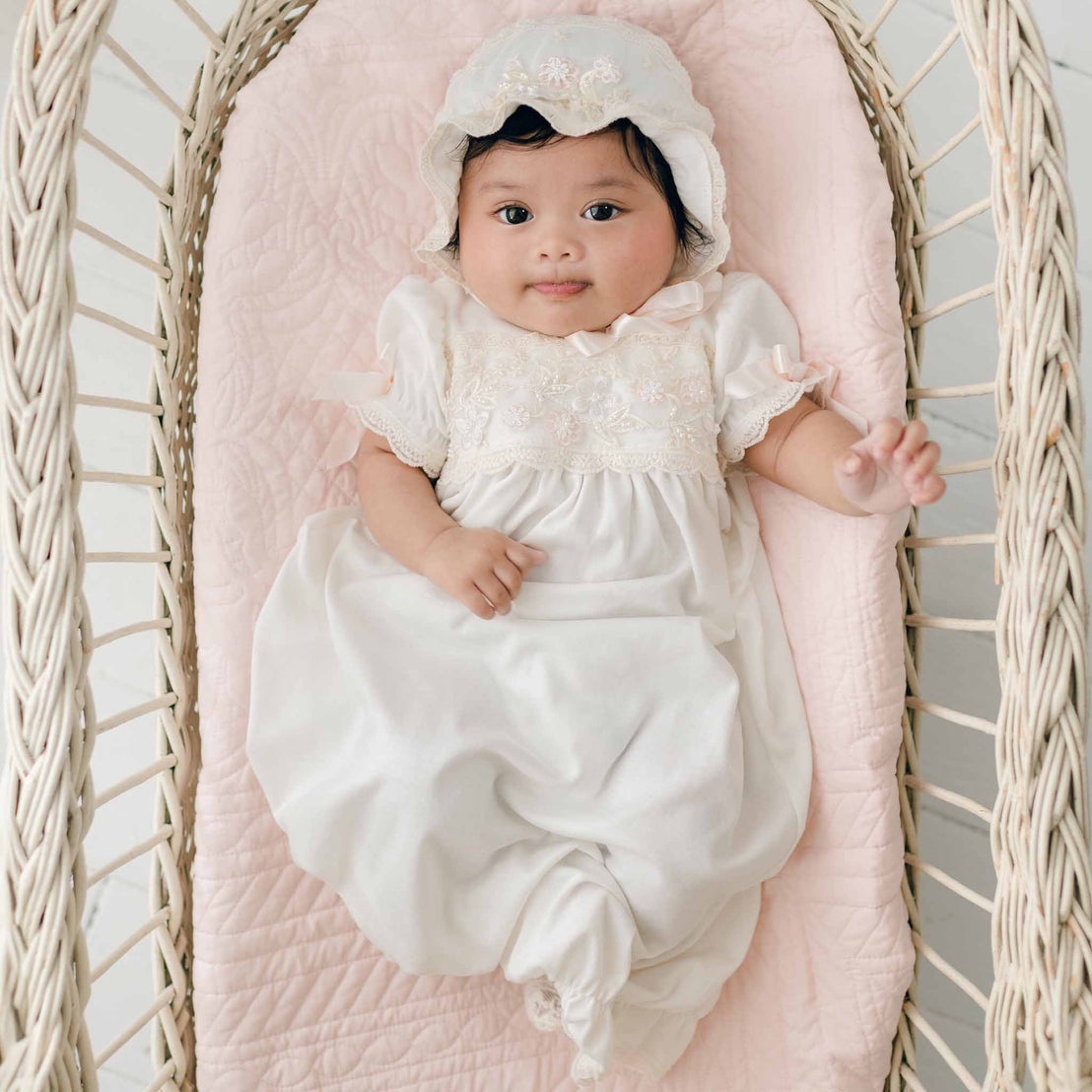 A baby wearing the Jessica Newborn Gown & Bonnet lies in a wicker bassinet lined with a pink quilted pad. The baby has a slight smile and one hand raised.