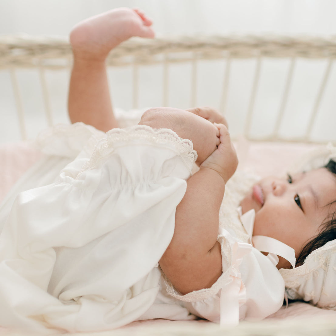 A baby wearing Jessica Newborn Gown & Bonnet lies in a crib, holding both feet in the air.