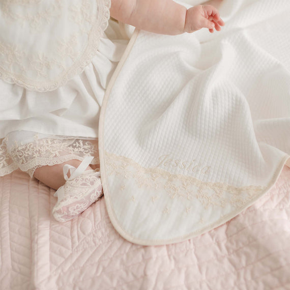 A baby dressed in white lace clothing sits on a pale pink quilted blanket, partially covered by a Jessica Personalized Blanket. The baby’s leg and foot, wearing a lace shoe, are visible—a perfect personalized baby gift.