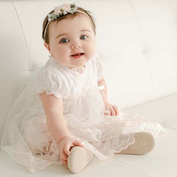 A baby in a Joli Romper Dress with embroidered netting lace and a floral headband sits on a white couch, looking forward with a slight smile.