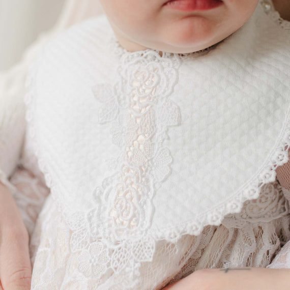 Close-up of a baby wearing a white textured cotton and lace bib with pink ribbon detail