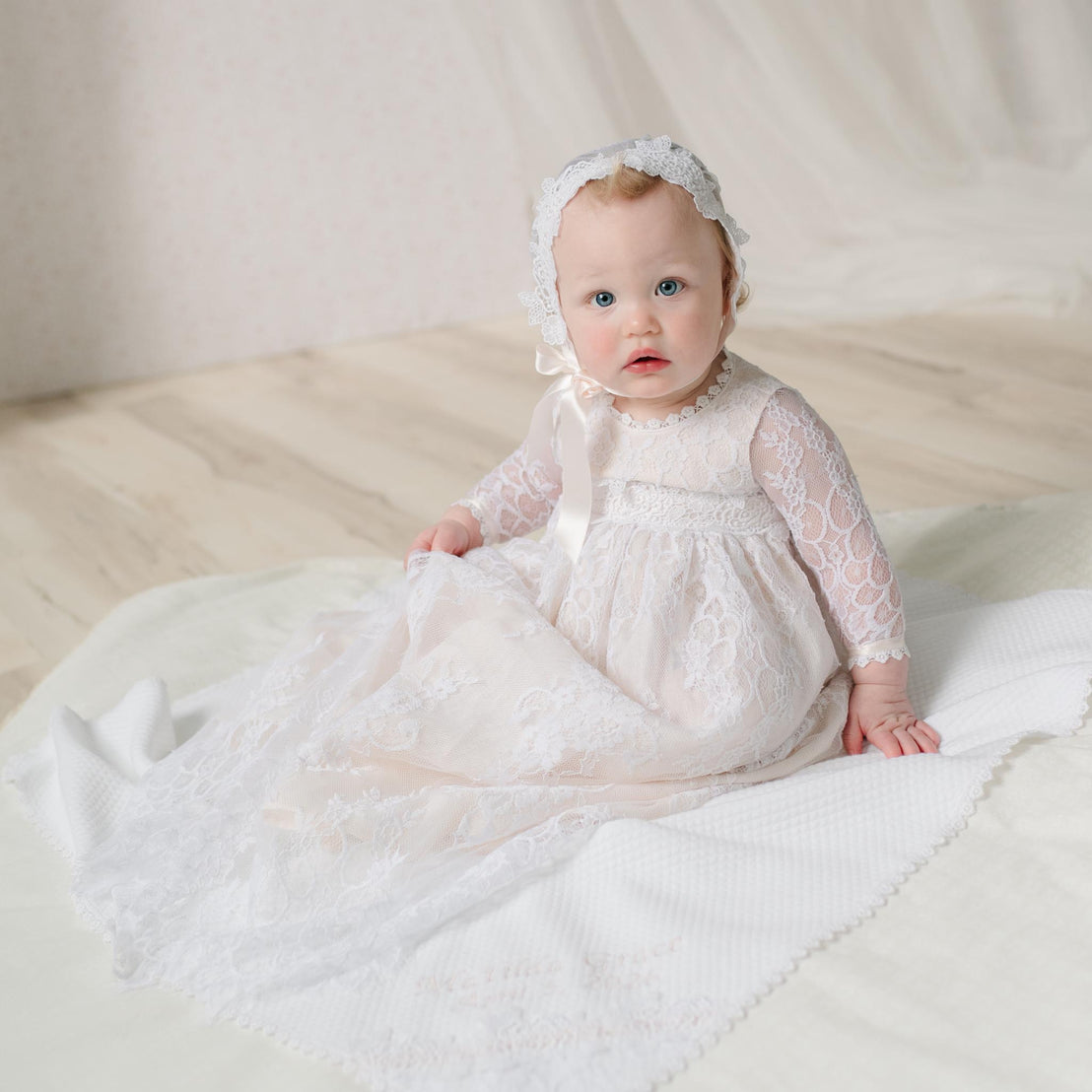 Baby in a white lace dress and bonnet sitting on a white blanket.