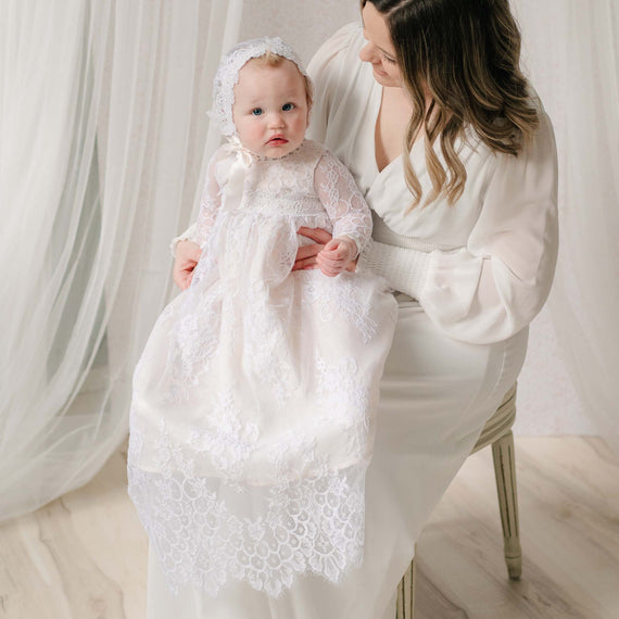 Mom sitting holding a baby dressed in a white lace gown and bonnet against a white curtain background.
