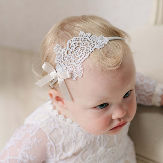 Baby wearing a white lace headband with a pink bow, sitting on a beige couch.