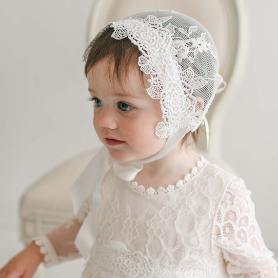 A toddler wearing a white lace Christening gown and a matching Juliette Lace Bonnet sits in front of a light-colored chair. The child has short brown hair and blue eyes, and the lace bonnet features a detailed floral pattern. The background is softly blurred.