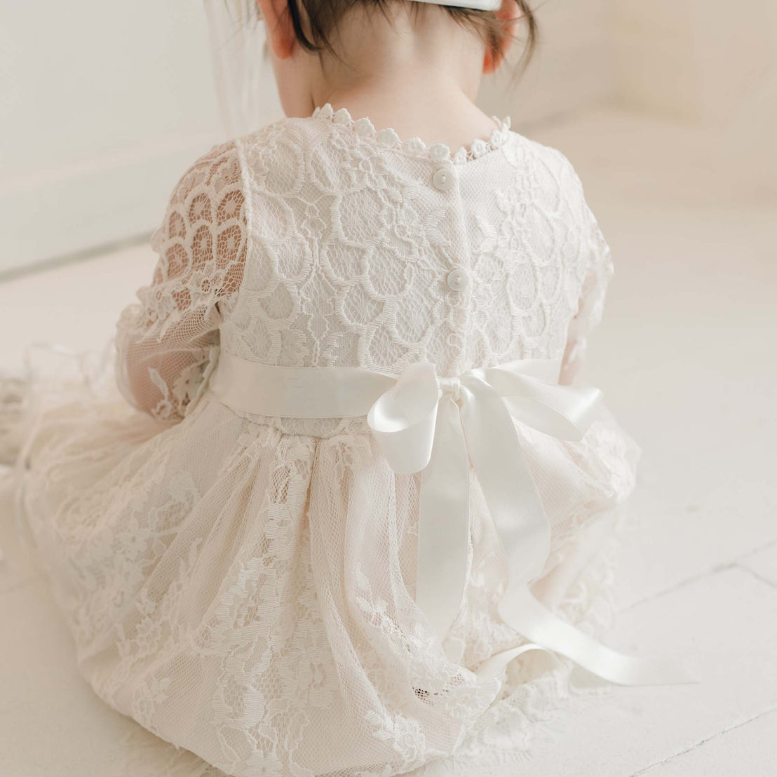 A young child with dark hair is sitting with their back to the camera, wearing the Juliette Romper Dress. This white, lacy handmade dress features intricate floral lace patterns and pearl-like buttons along the back, with a silk ribbon tie in a bow at the back.