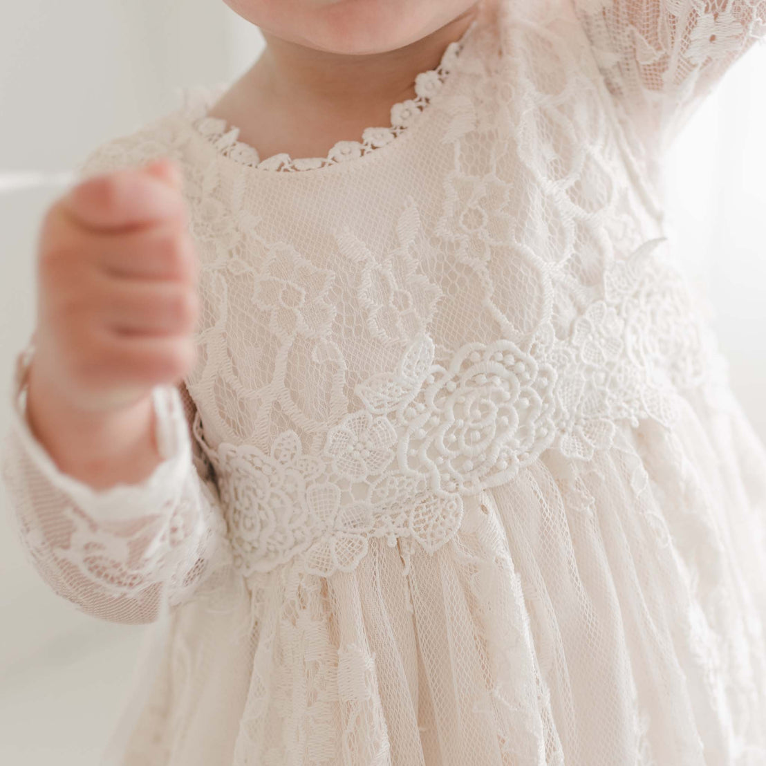 Close-up of a young child wearing the Juliette Romper Dress, adorned with intricate floral lace and a decorative waistband. The child's face is not visible, and their arms are raised slightly, holding a small object. A soft silk ribbon tie adds an elegant touch to the softly lit and blurred background.