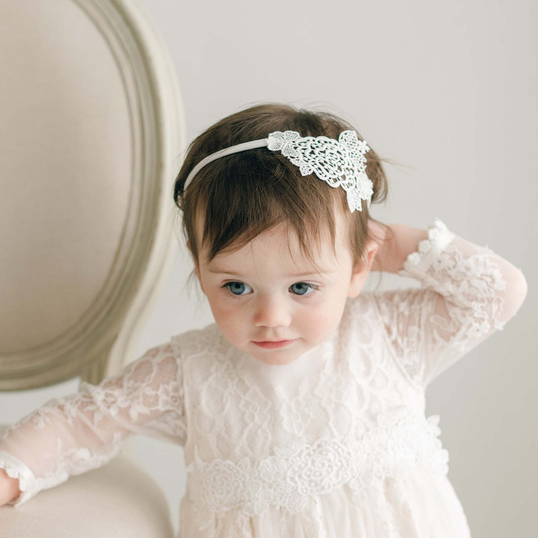 A young child with dark hair is wearing a white lace dress adorned with light ivory rose lace, paired with the elegant Juliette Lace Headband while seated. The child has one arm raised behind their head and is looking slightly downward. A light-colored chair with a curved back is visible in the background.