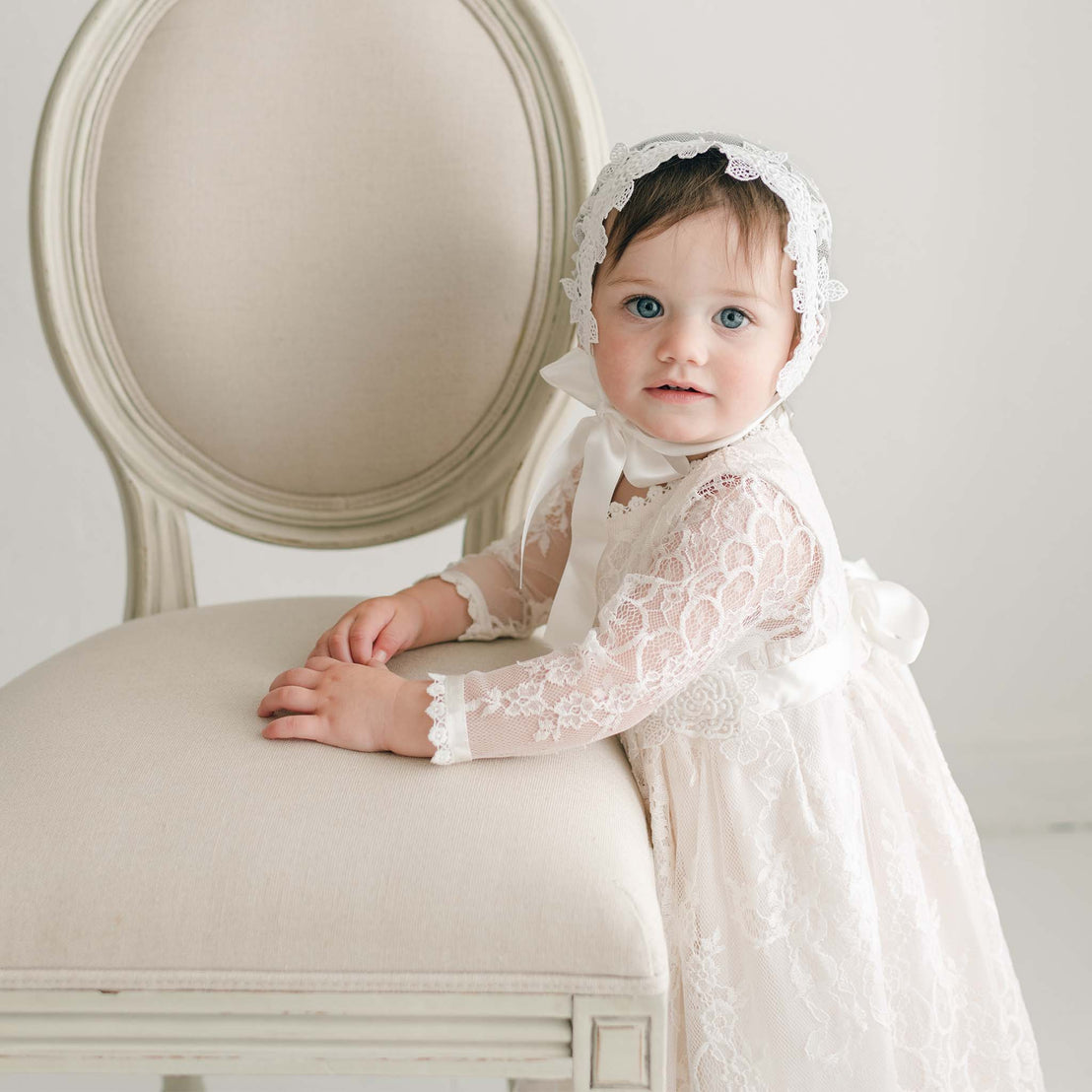 A baby, dressed in the Juliette Romper Dress featuring floral lace and adorned with a silk ribbon tie, stands while holding onto a light-colored upholstered chair. The baby has blue eyes and dark hair, with a neutral and minimally detailed background adding focus to the charming scene.