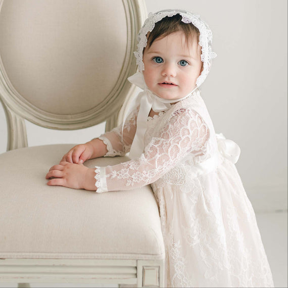 A baby, dressed in the Juliette Romper Dress featuring floral lace and adorned with a silk ribbon tie, stands while holding onto a light-colored upholstered chair. The baby has blue eyes and dark hair, with a neutral and minimally detailed background adding focus to the charming scene.