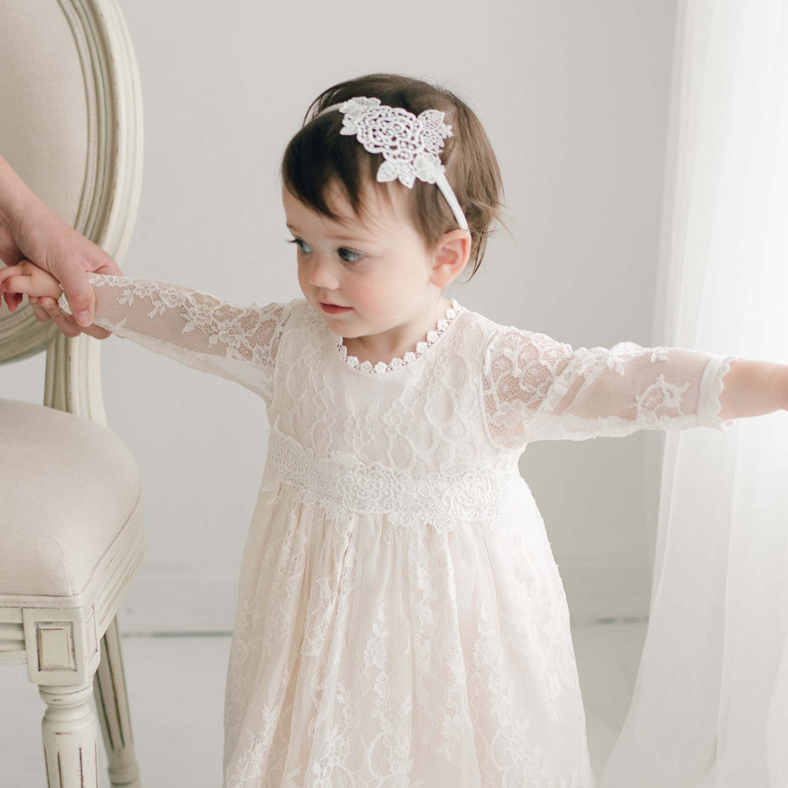 A toddler with short brown hair wearing the Juliette Romper Dress and a lace headband holds an adult's hand. She stands beside a beige upholstered chair, her other arm stretched out to the side, looking focused on something in front of her.