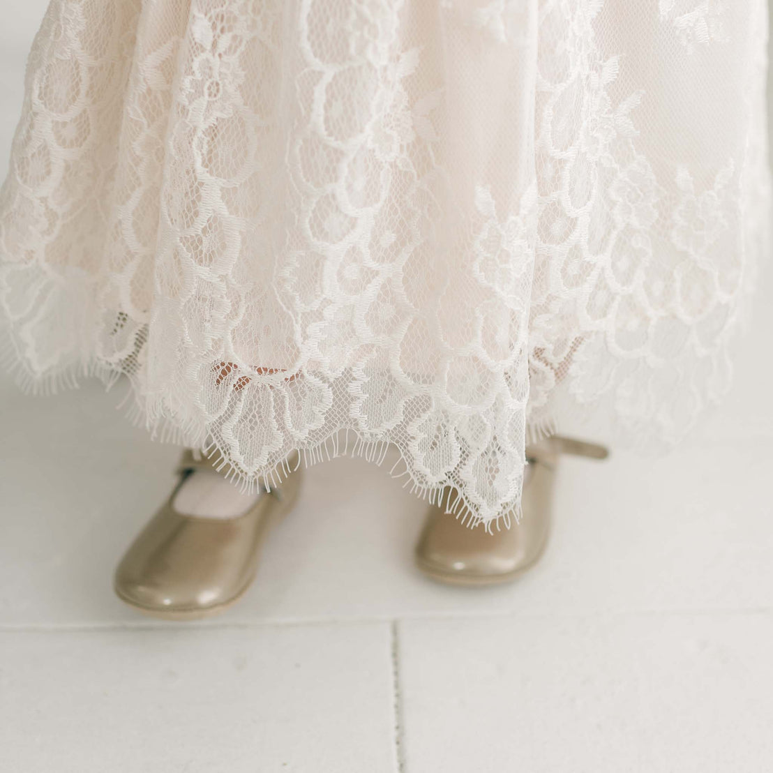 A child is wearing the Juliette Romper Dress, a cream-colored lace outfit with scalloped edges that extend down over their feet, paired with gold-colored strap shoes. The dress features a delicate silk ribbon tie at the waist, adding an extra touch of elegance. The shoes are positioned on a white surface.