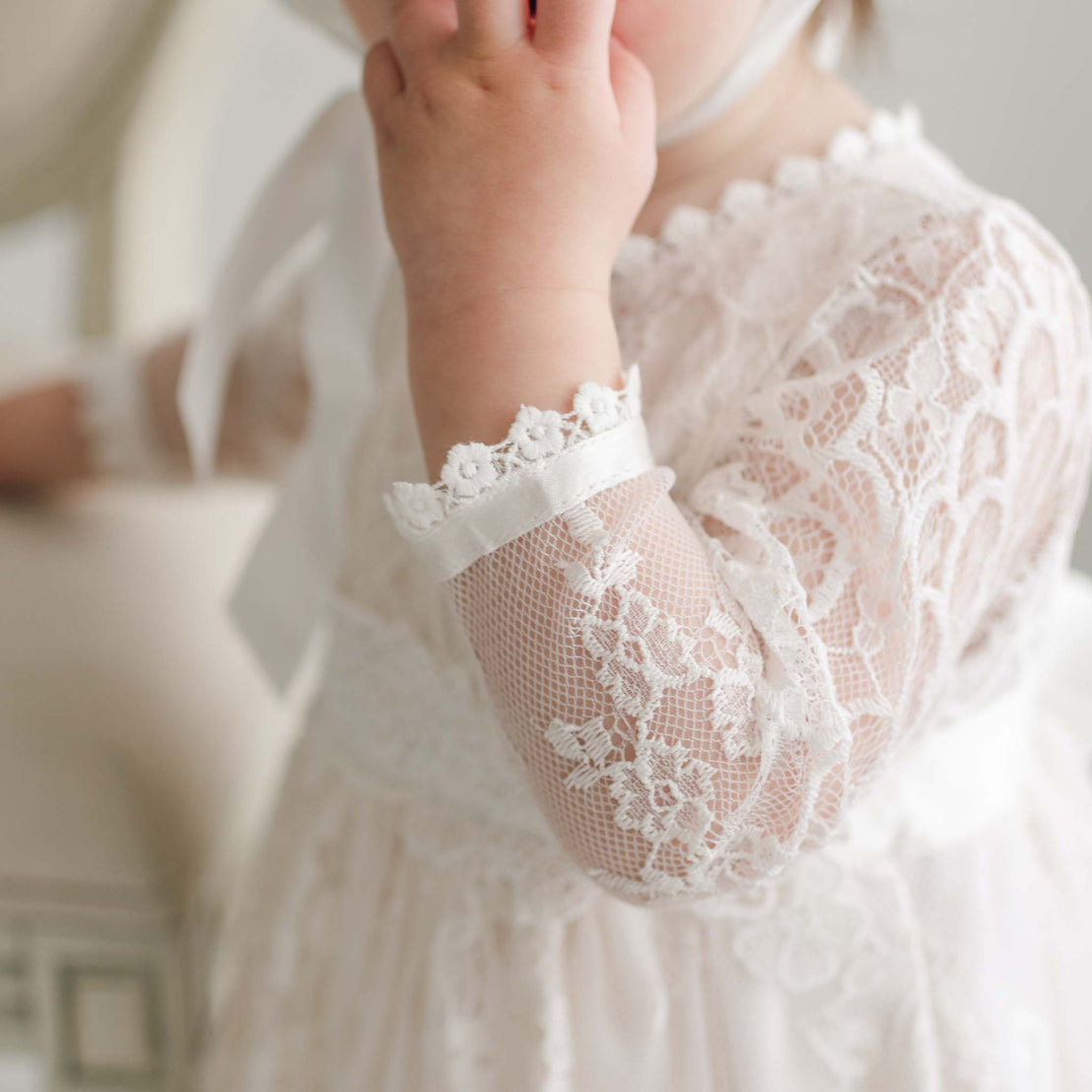 Close-up of a child in the Juliette Romper Dress with long sleeves, their hand partially covering their mouth. The romper features intricate floral lace patterns and detailed silk ribbon ties. The background is out of focus, emphasizing the delicate details of the clothing.