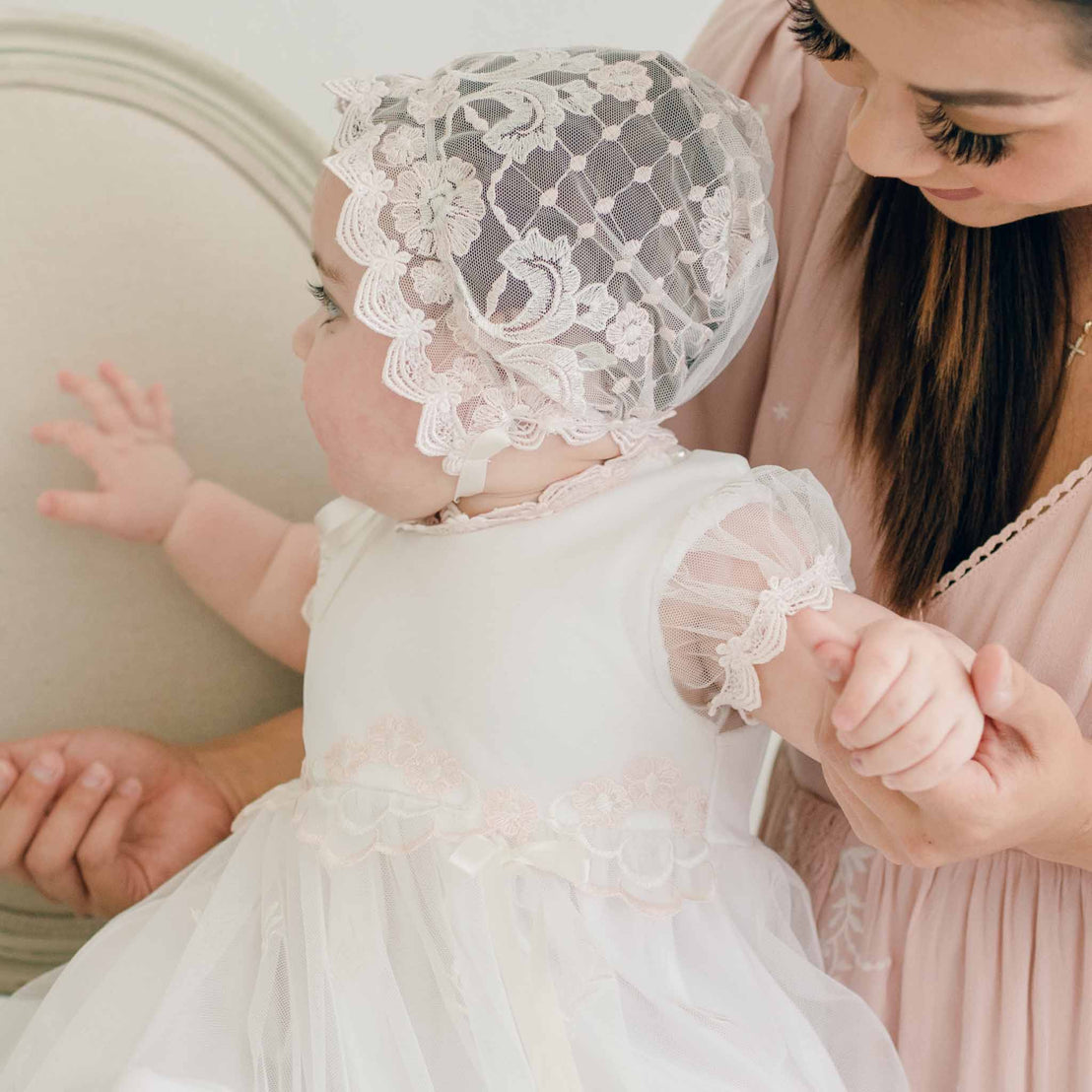 Baby in a lace bonnet and dress held by a woman in a soft pink dress.