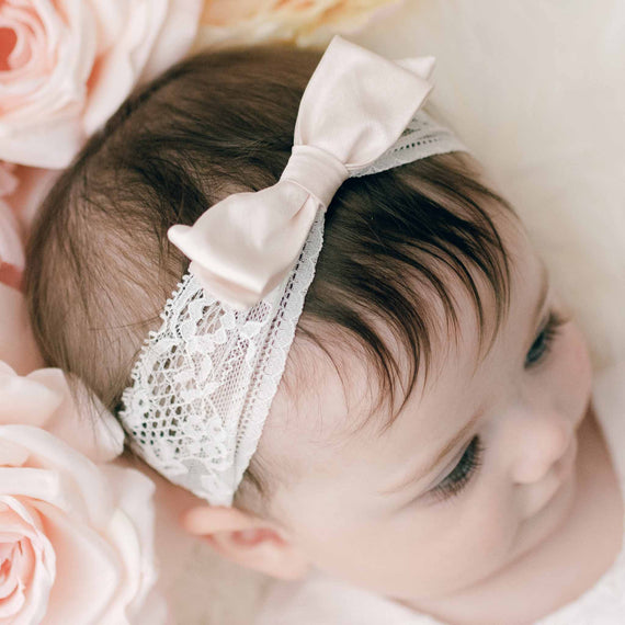 A baby with brown hair wears the Victoria Lace Headband, crafted from luxurious pink champagne silk, and is surrounded by pink roses. The baby gazes to the side, partially visible. This delicate headband, handmade in the USA, adds a touch of elegance to the scene.