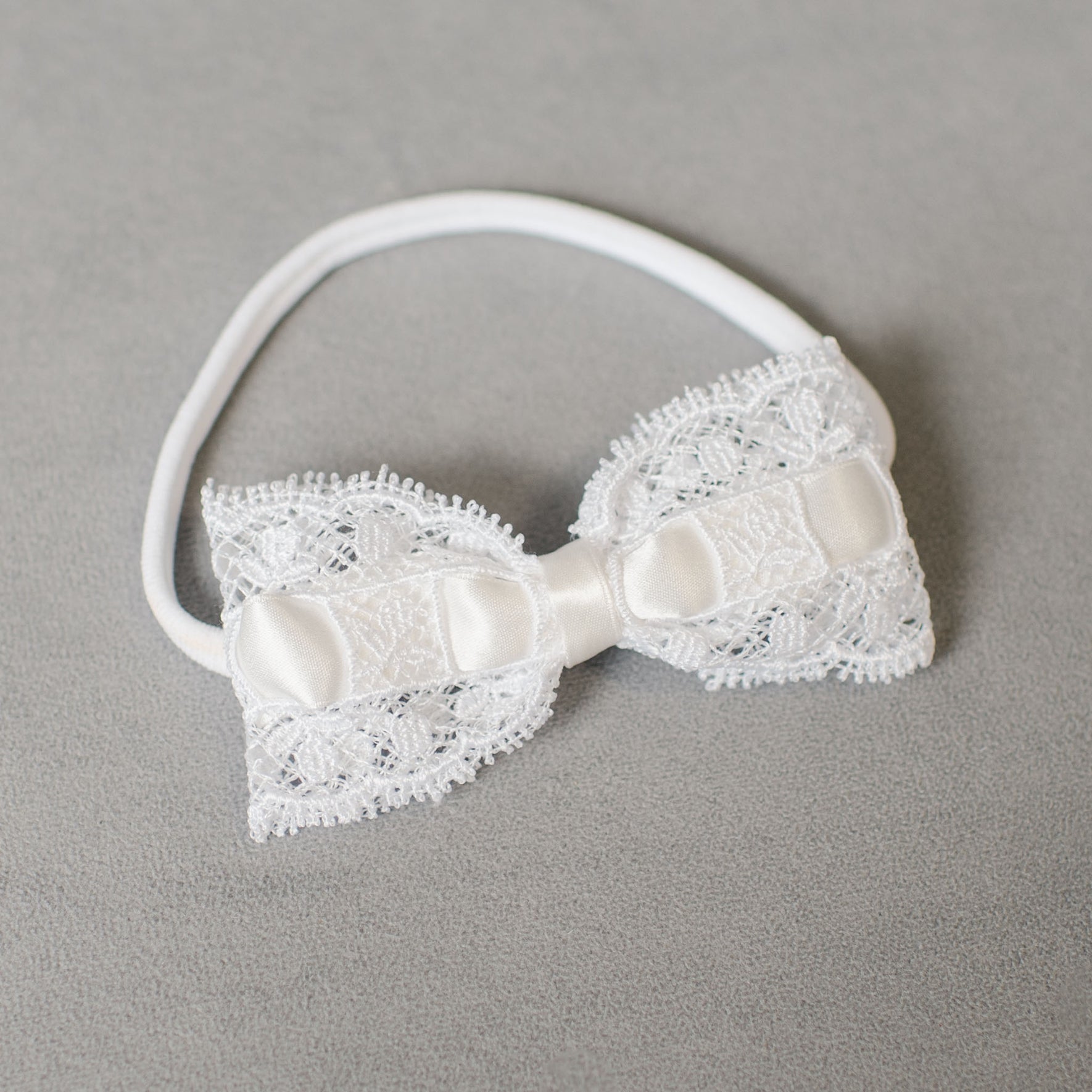 A white lace headband with a decorative bow crafted from lace and satin lies on a gray surface, elegantly paired with an Aria Christening gown. The bow showcases detailed lace work and a sleek satin center, making it an ideal baptism accessory included in the Aria Accessory Bundle - 15%.