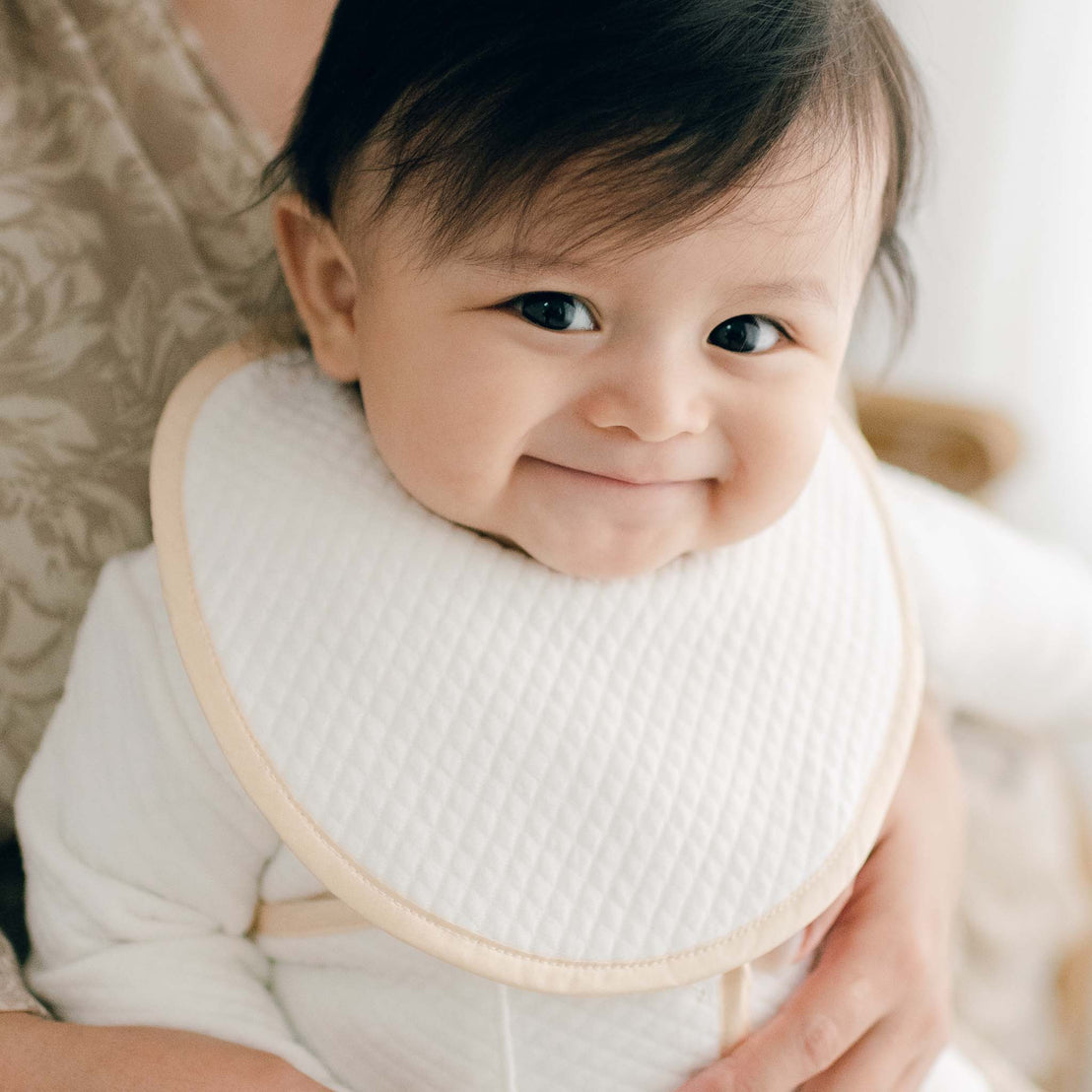 Baby boy smiling and wearing the Liam Boys Bib crafted from soft quilted cotton in ivory and detailed with a silk champagne trim